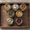 Top-down editorial flat-lay of seven mason jars on a walnut tray, each containing different asset-class objects representing portfolio diversification